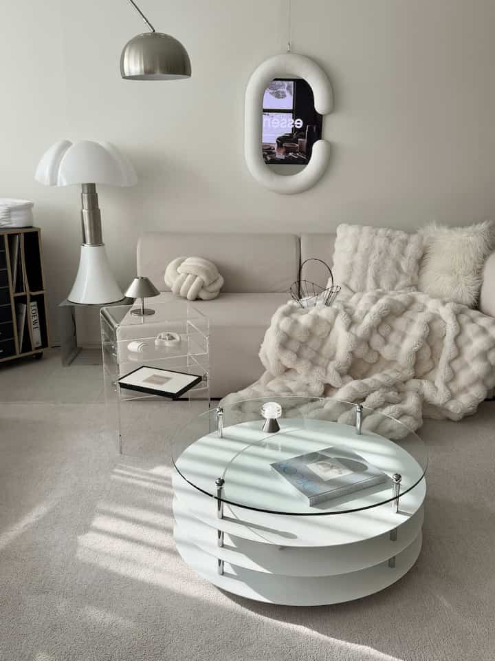 ナチュラルな色調のリビング、一面にベージュのファブリックソファと丸いガラスコーヒーテーブル、ファーのブランケットとシルバーの照明が配置された雰囲気のあるインテリア