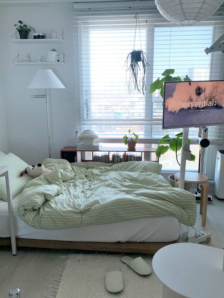 A clean bedroom with white and wood tones, featuring a bed, blinds, and plants in a natural modern atmosphere