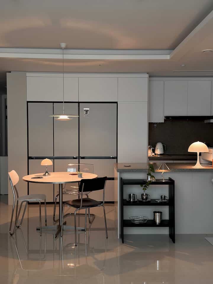 ホワイトとグレーのトーンのキッチン、丸いダイニングテーブルと椅子、ペンダントライトと棚が配置された雰囲気のあるインテリア