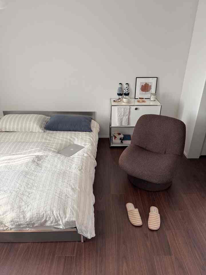 Minimal bedroom with white walls and bed, brown wooden floor, and brown armchair