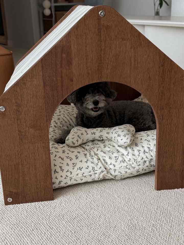 ナチュラルな木目調のペットハウスと白いフロアラグの上に、花柄のクッションでくつろぐグレーの犬がいる温かみのある空間