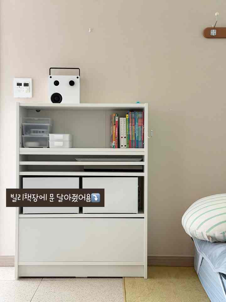 明るく落ち着いたベージュの壁を背景に、白い本棚と白いBluetoothスピーカーが配されたモダンでミニマルなインテリア空間