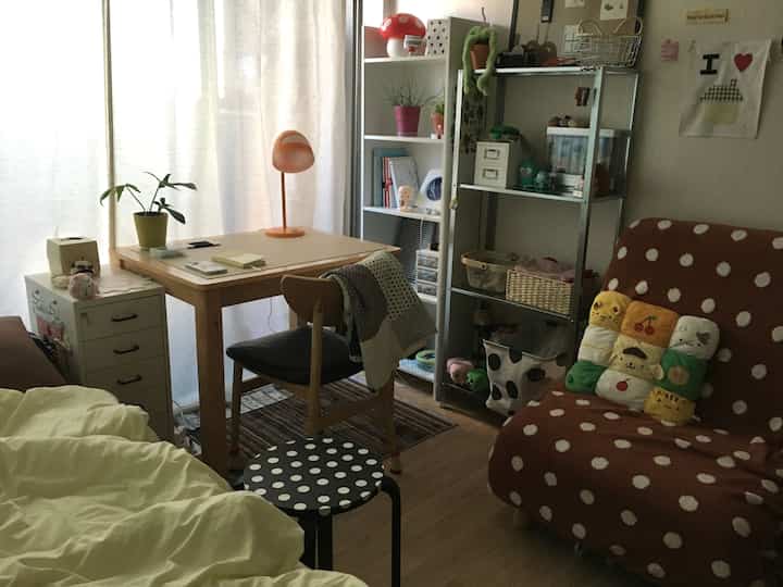 明るいベージュと茶色のトーンのコンパクトなワンルームリビング、机と椅子、ソファ、収納棚が配置された居心地の良い空間