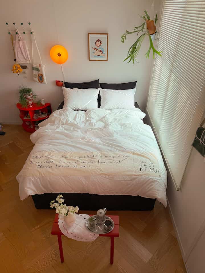 A bright white and wood-toned bedroom featuring a large bed centerpiece and red nightstand, creating a simple and natural atmosphere