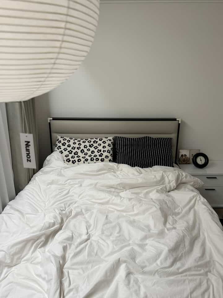 White-toned bedroom featuring a large central bed with white bedding, simple modern style with a nightstand and clock on the right