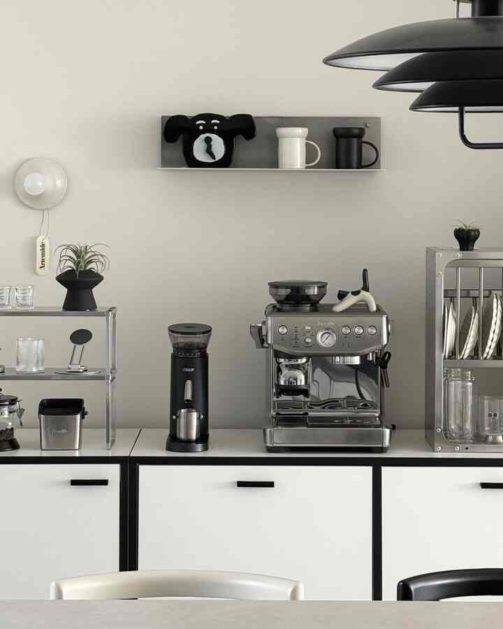 モノトーンとブラックが調和するキッチンのホームカフェ、メタル棚とコーヒーマシン、壁棚には犬の時計とマグカップが配置された印象的な空間