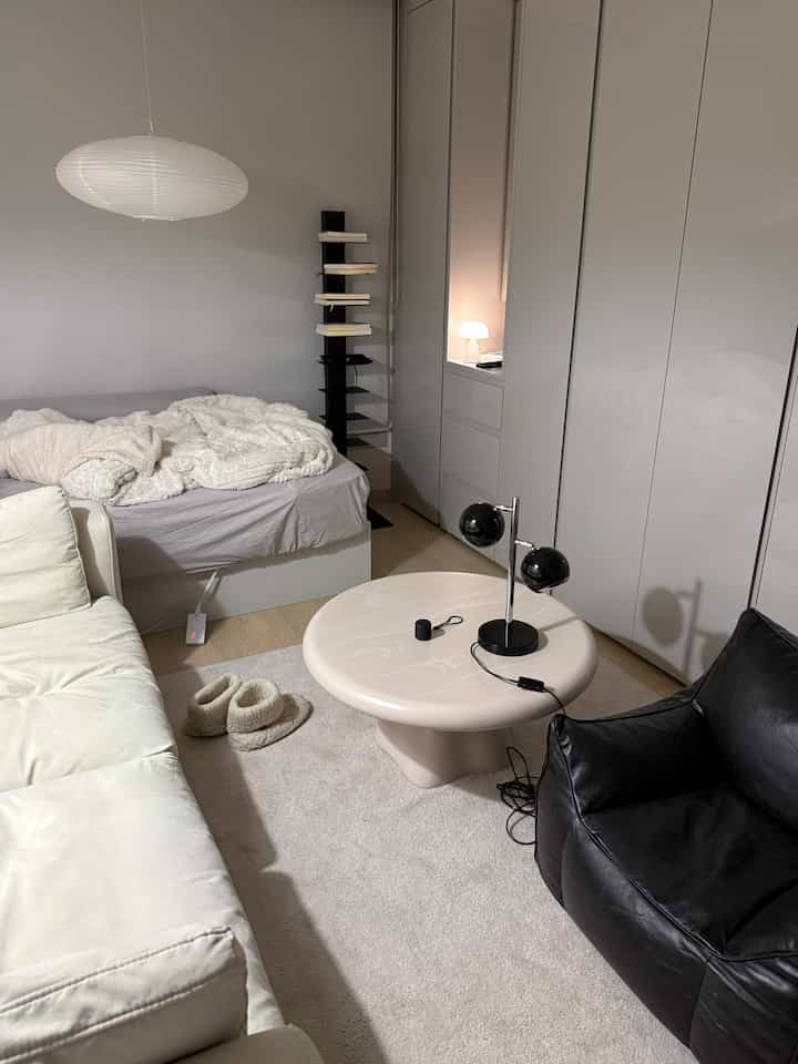 ナチュラルな色調の寝室とリビングスペース、ベージュのコーヒーテーブルとクリーム色のソファ、黒いビーズクッションが配置された落ち着いた空間