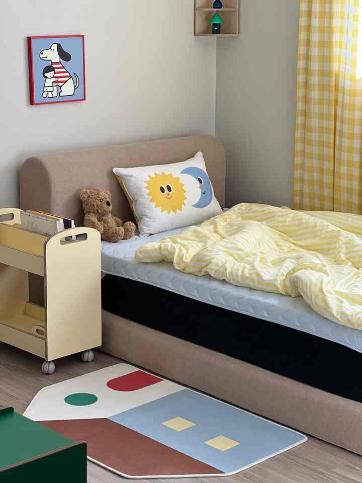 明るい黄色のカーテンと掛け布団カバーがある子供部屋、ベッドと絵本カート、かわいいラグが配置された雰囲気のあるインテリア