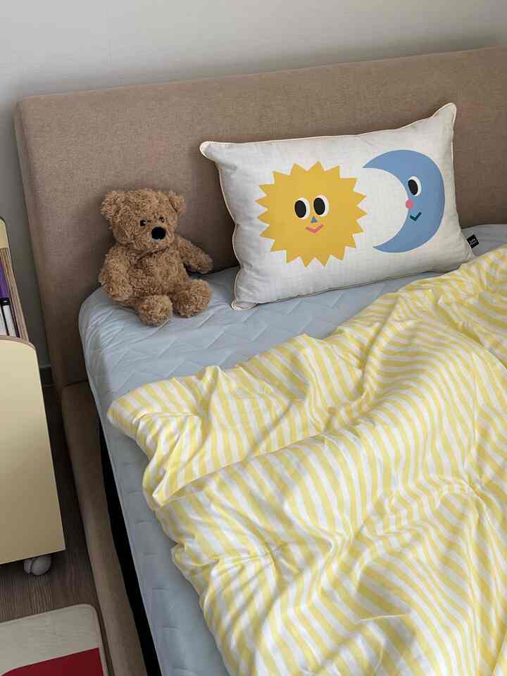 ベージュのヘッドボードと黄色のストライプの寝具、太陽と月の柄の枕、クマのぬいぐるみが置かれた子供部屋の温かみのある空間