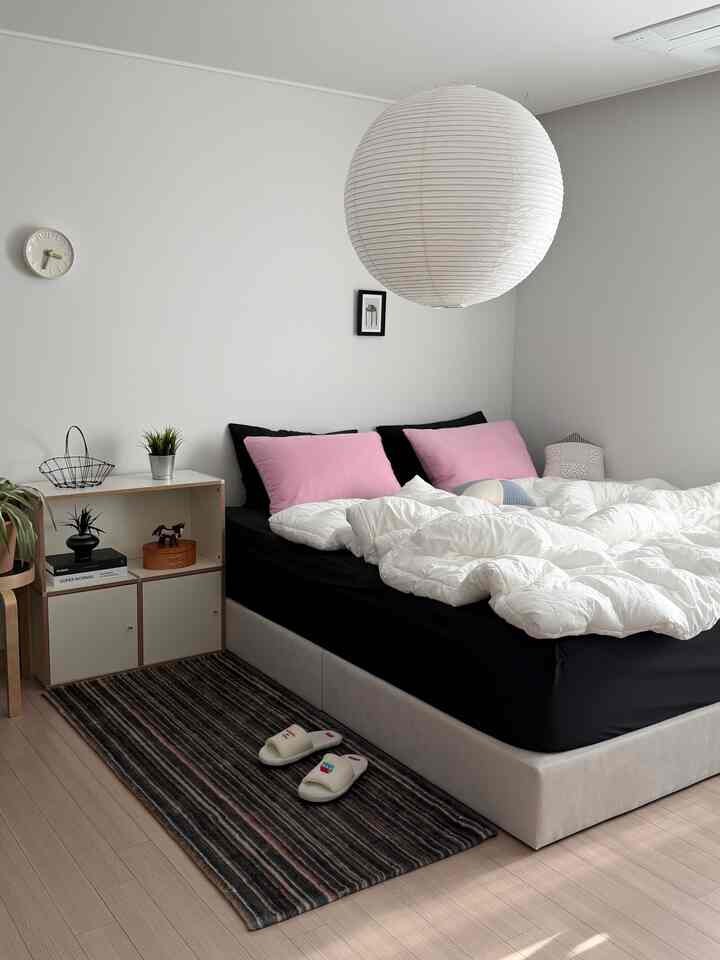 白い壁と天井の寝室、中央にベージュのベッドとピンクの枕があり、左側にモジュール式の本棚が配置された清潔感のあるインテリア