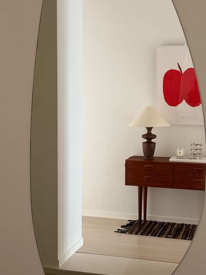 白色調の玄関、木製コンソールテーブルとテーブルランプ、赤いリンゴの絵が配置されたミニマルな空間
