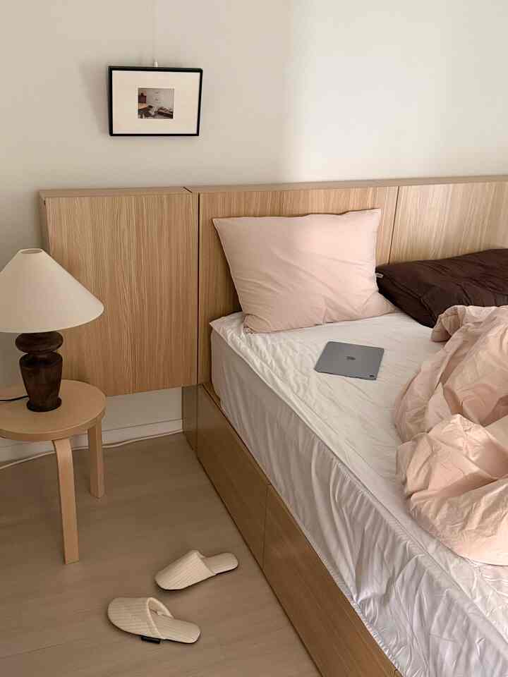 柔らかな木目調と白を基調とした寝室、ベッドとナイトテーブル、テーブルランプが配置された落ち着いたミニマルなインテリア