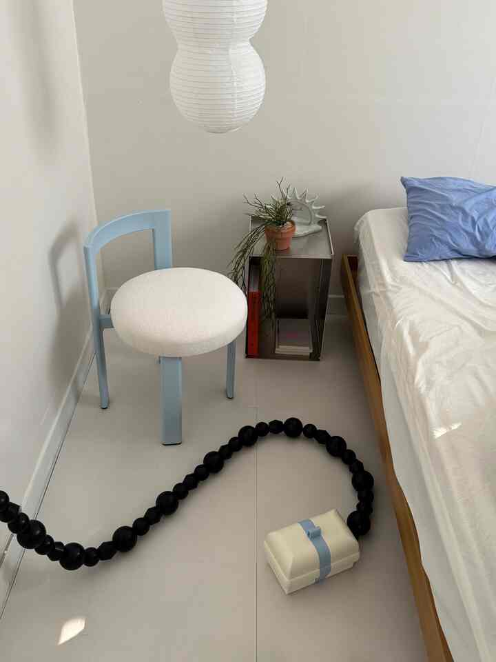 白い壁と床、透明感のある照明があるミニマルでナチュラルな寝室、ライトブルーのスツールと木製ベッドフレームが調和したインテリア。