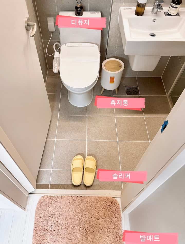 白と茶色のタイル床が印象的なバスルーム、トイレと洗面台、ピンクのバスマットとベージュのスリッパが配置された清潔感のある空間
