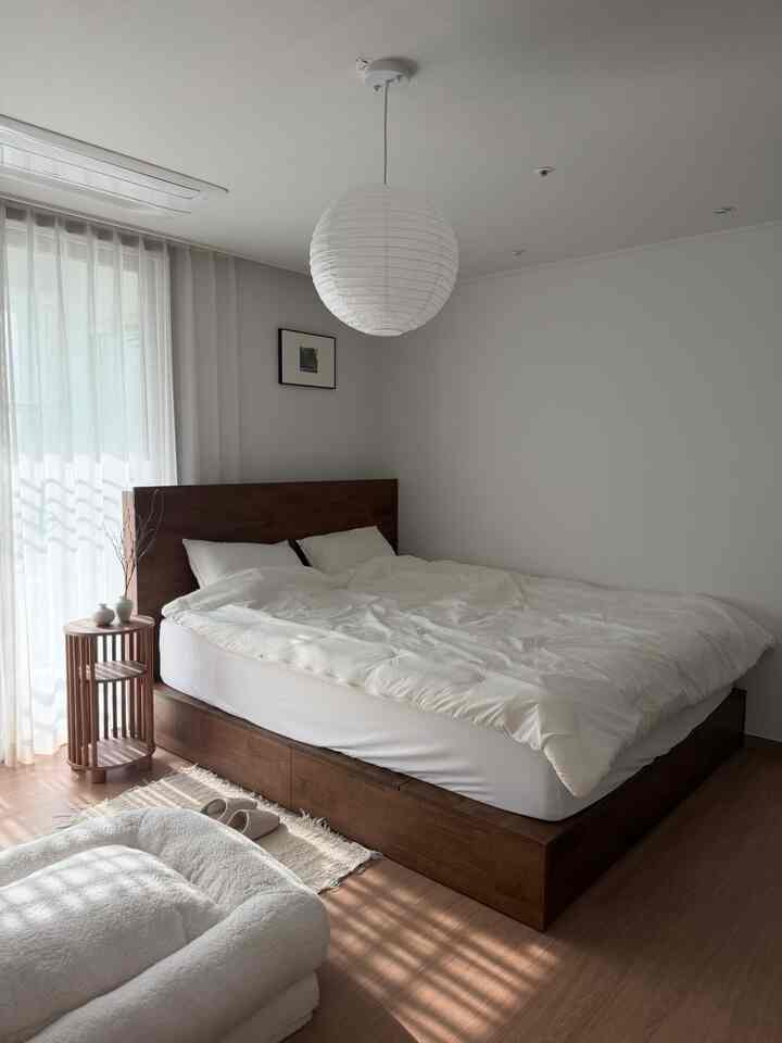 ホワイトとウッドトーンが調和するモダンな寝室、大きなベッドとナイトテーブル、カーテン、フロアチェアが配置された雰囲気のあるインテリア