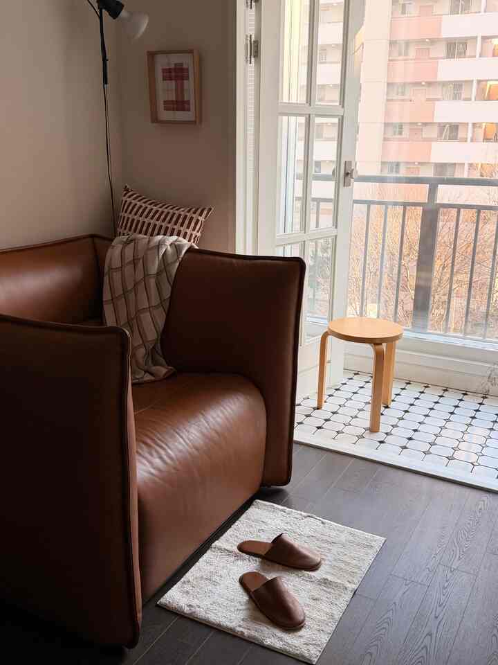 ブラウンのレザーソファと小さなマットが置かれたナチュラルトーンのリビング、広いガラス窓からバルコニーが見える印象的な空間