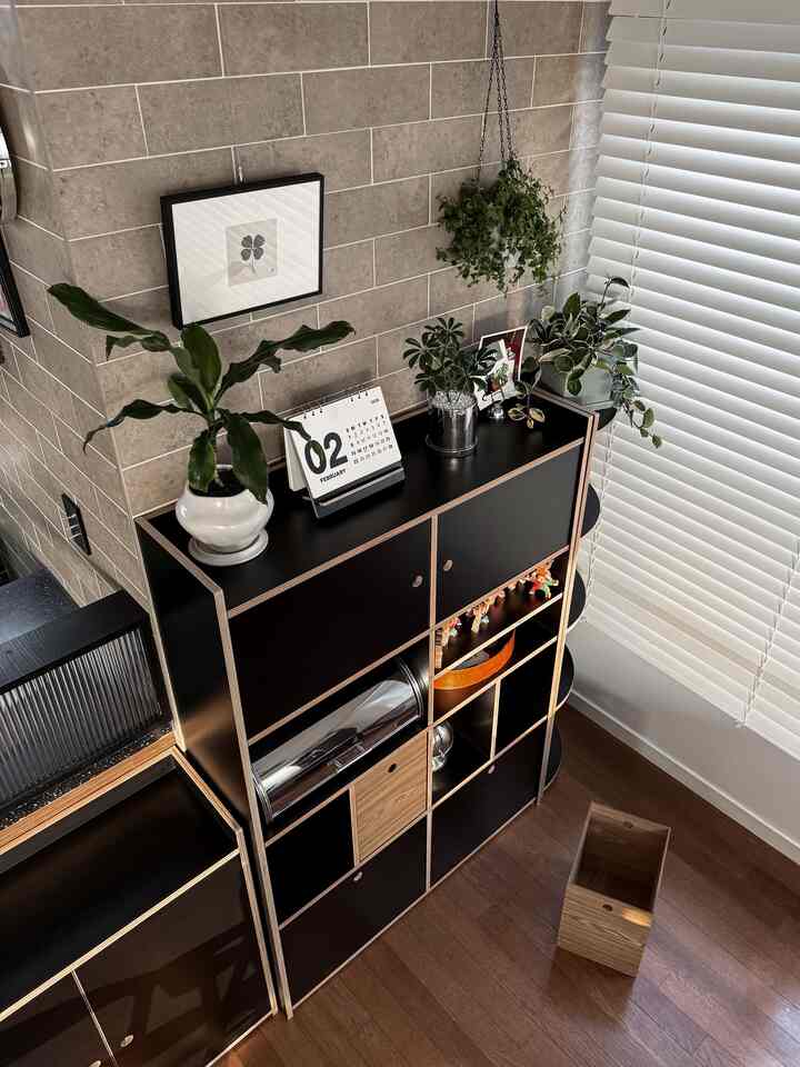 グレーのタイル壁と黒の木目調収納キャビネットが配置されたコーナースペース、吊り下げ植物と白いブラインドが調和する清潔感のあるモダンなインテリア