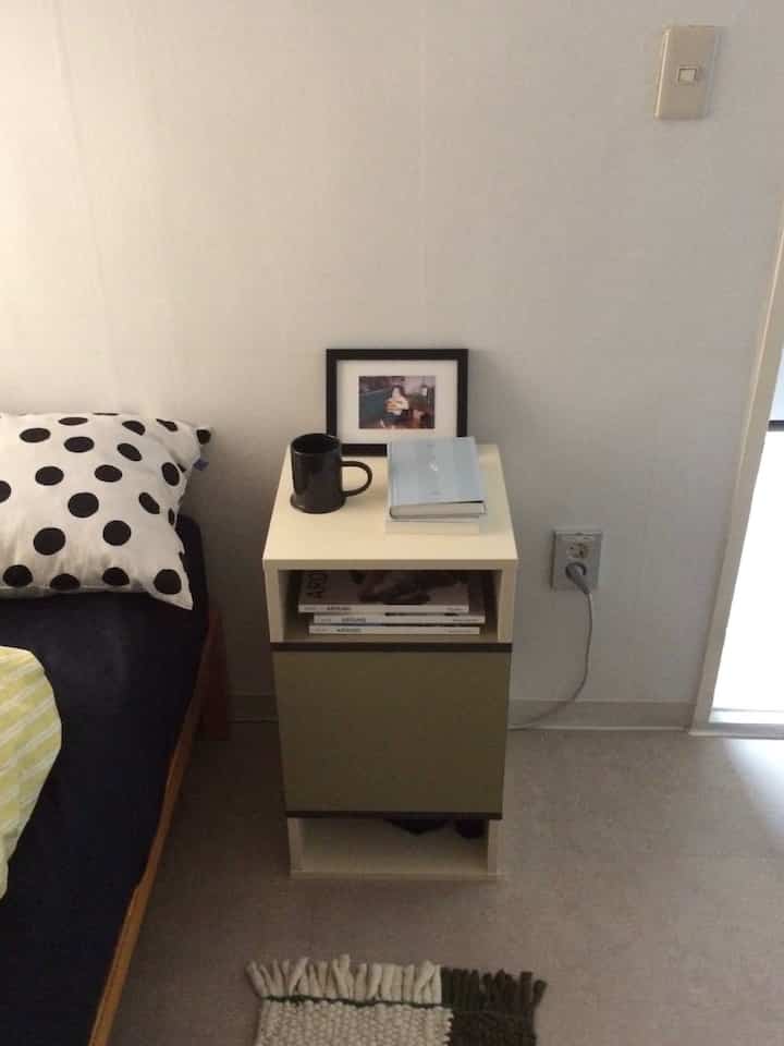 白い壁とネイビーベッドがある小さな寝室、ナイトテーブルの上にカップと本が置かれた雰囲気のあるインテリア