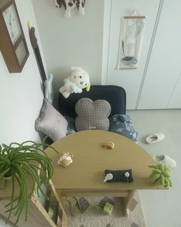 ナチュラルな色調のコンパクトな部屋、小さな丸テーブルとネイビーのアームチェア、クッションとぬいぐるみが置かれたシンプルな空間