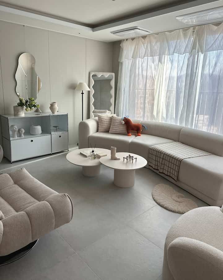 ナチュラルな色調のリビング、ベージュのソファと窓辺の白いカーテン、馬の形のクッションがアクセントの印象的な空間