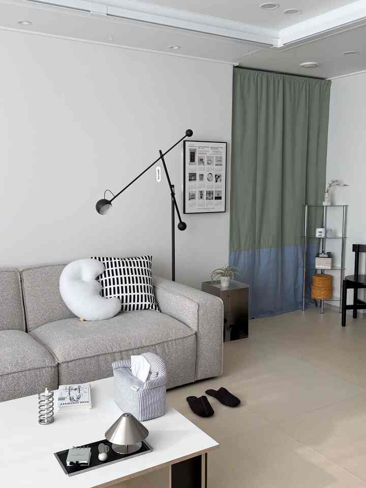 グレーのソファと白いコーヒーテーブルがあるシンプルでモダンなリビング、緑と青のカーテンとコーナーシェルフが配置された空間