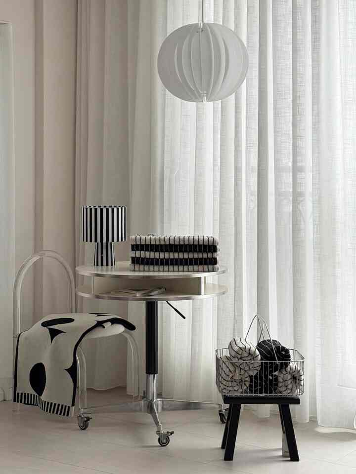 モノトーンのリビング、丸テーブルに折りたたまれたストライプのタオルとパターンのスローが置かれ、透明なアクリルのスツールと白いペンダントライトが配置された清潔感のある空間