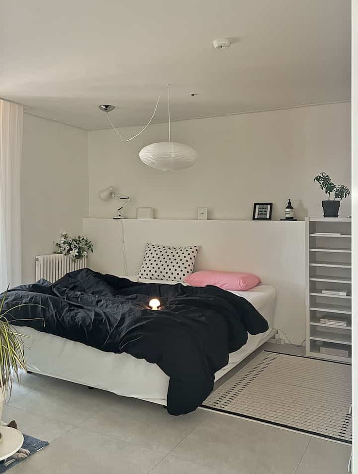 A white and black toned bedroom featuring a low bed frame, pillow covers, radiator, and lighting in a simple modern setting