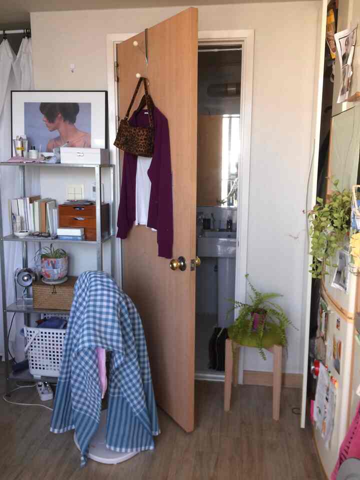 白い壁と木目調の床が特徴の小さなワンルーム、金属製の棚と緑のスツールに植物が置かれた雰囲気のあるインテリア
