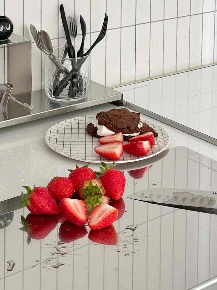 白いタイルの壁とステンレスのカウンターがあるキッチン、透明な容器に包丁やカトラリーが配置され、チョコレートクッキーとイチゴが皿とカウンターに置かれた清潔な空間