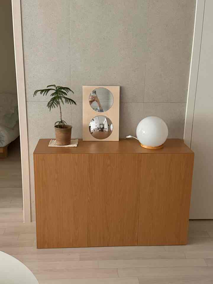 ナチュラルな木目調の玄関、木製キャビネットの上に鏡と植物、丸いムードランプが配置されたミニマルなインテリア