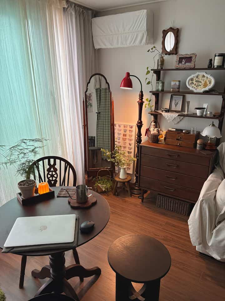 ナチュラルな木目調と赤い照明が調和したリビング、丸い木製のダイニングテーブルとスツール、柔らかなカーテンが温かみのある雰囲気のインテリア