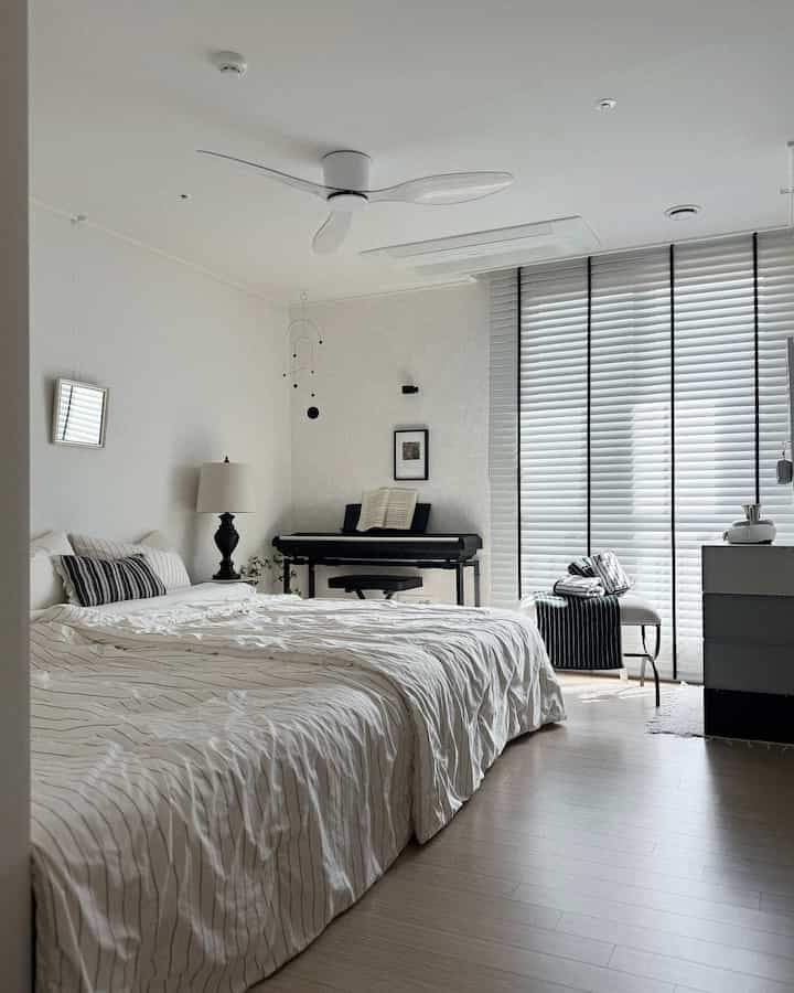 白とベージュの色調の寝室、広いベッドとブラインド、ピアノとスツールが配置された雰囲気のある空間