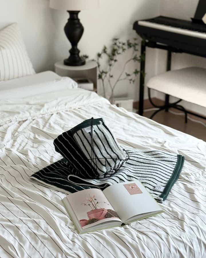 白と緑のトーンの寝室、すっきりとしたベッドの上にストライプのタオルと開かれた本が置かれた落ち着いた雰囲気のインテリア