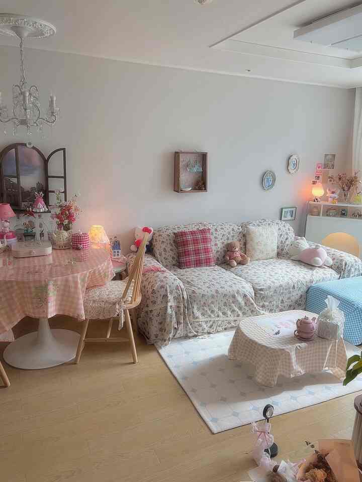 淡い白の壁と木目調の床のリビング、花柄ソファとピンクのギンガムチェックのテーブルクロスが配置された温かみのある空間