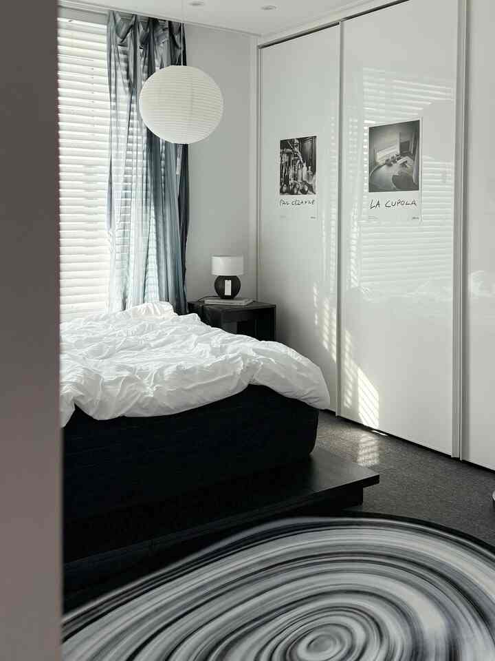 グレートーンのカーテンと黒いベッドがあるモノトーンの寝室、しわのある白い寝具と丸いテーブルランプが配置された印象的な空間