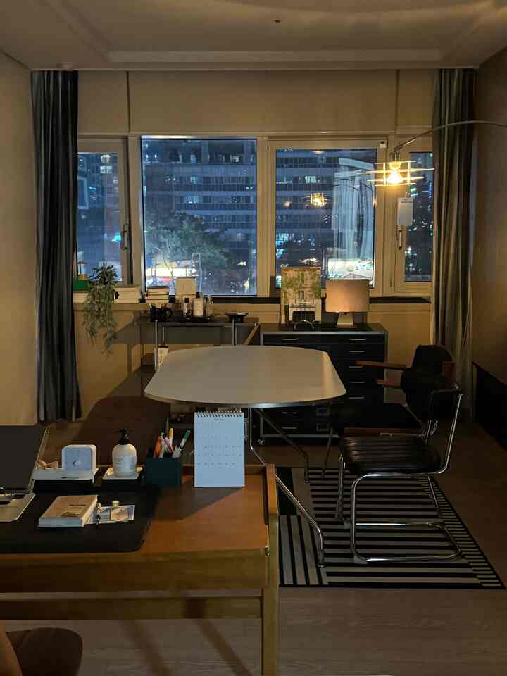 Dimly lit living room featuring mid-century modern desks and chairs centrally arranged, with large windows revealing city night views