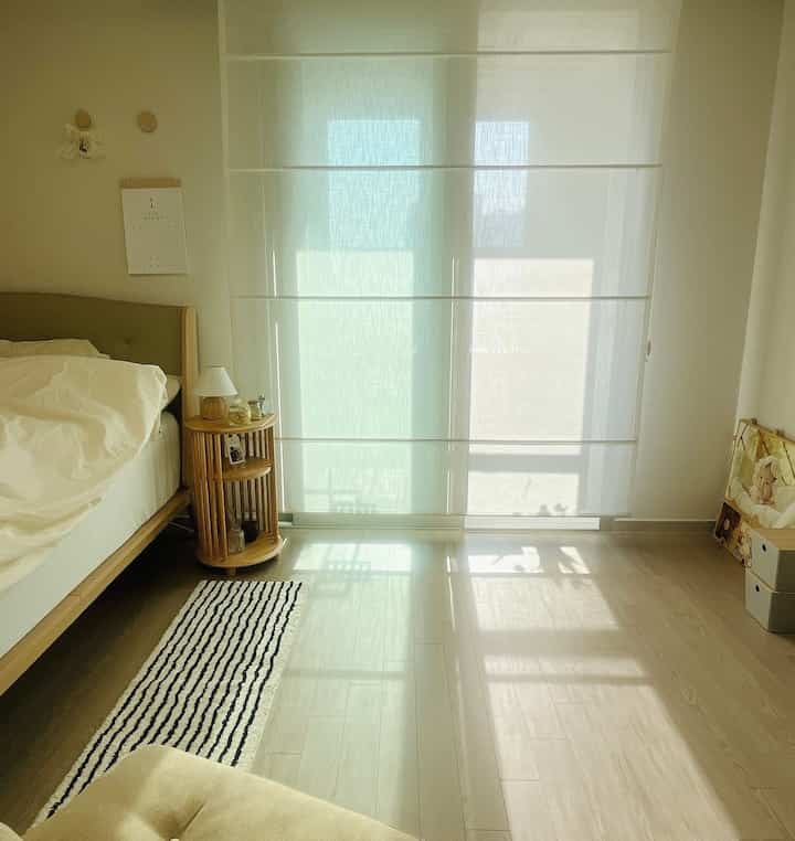 ホワイトとブラウンの色調の寝室、ローマンシェードのカーテンとベッド、ナイトテーブルが配置された落ち着いた空間