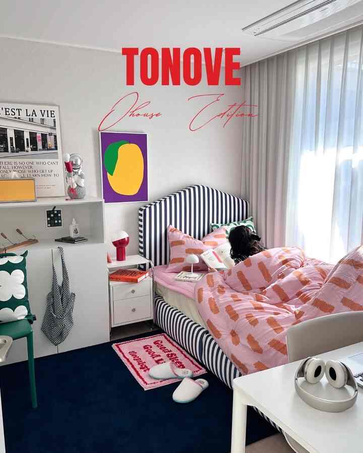 白い壁とカーテン、ネイビーストライプのベッドフレームとピンクの布団が配置されたコンパクトな寝室