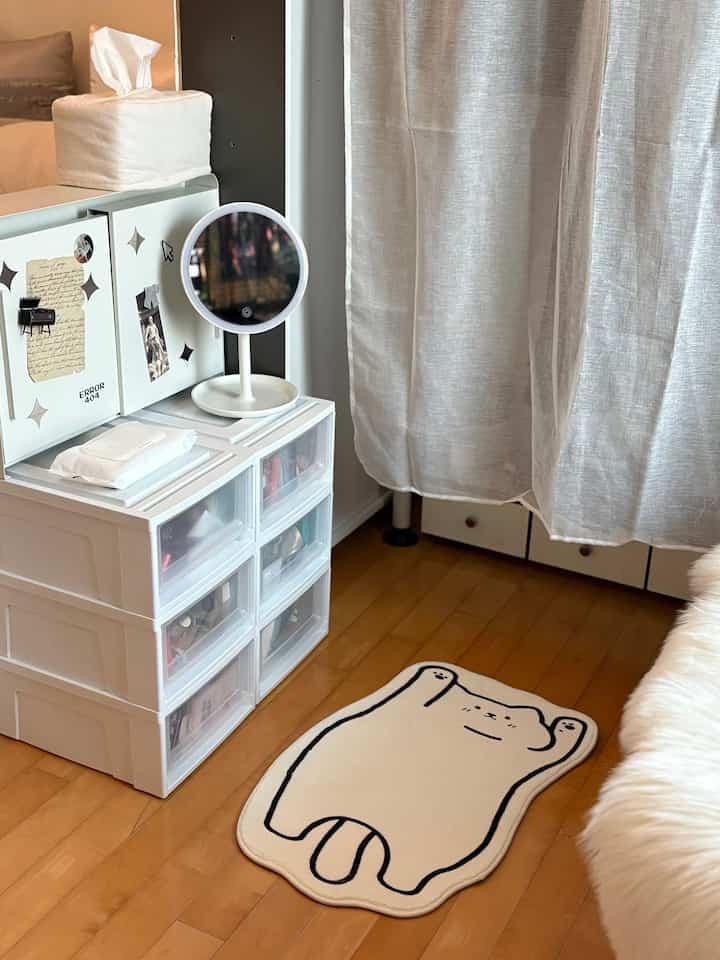 白と木目調の調和が取れた小さな部屋のコーナー、収納棚とLEDミラー、かわいいアクセントマットが配置された印象的な空間