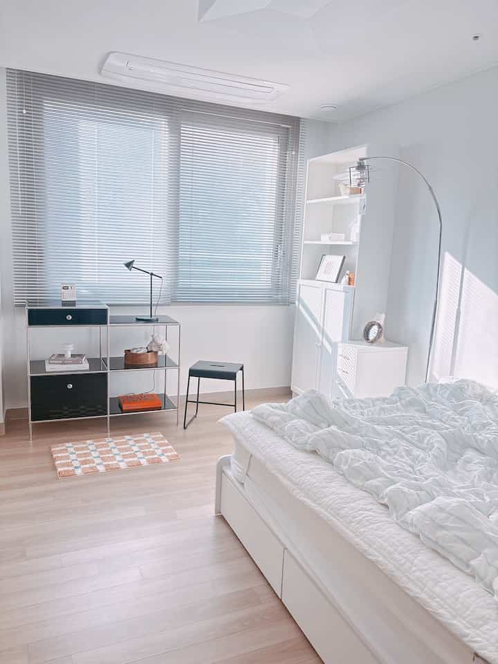 Bright white and wood-toned bedroom featuring blinds, cabinets, and a floor lamp, styled in a clean modern aesthetic
