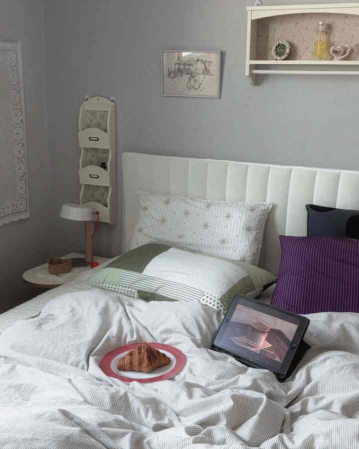 グレーの壁と白いベッドヘッドボードがある6畳の寝室、様々なクッションと寝具が配置された雰囲気のある空間