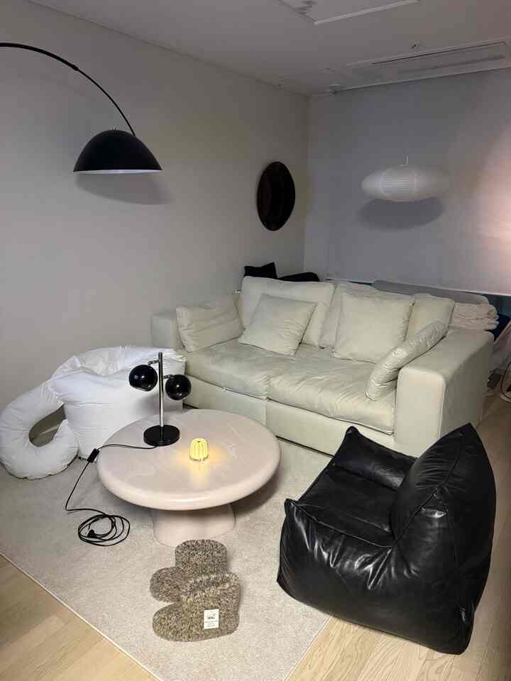 白とクリーム色のトーンが調和したワンルームリビング、クリーム色のソファと黒のビーンバッグ、丸いコーヒーテーブルが配置されたミニマルな空間