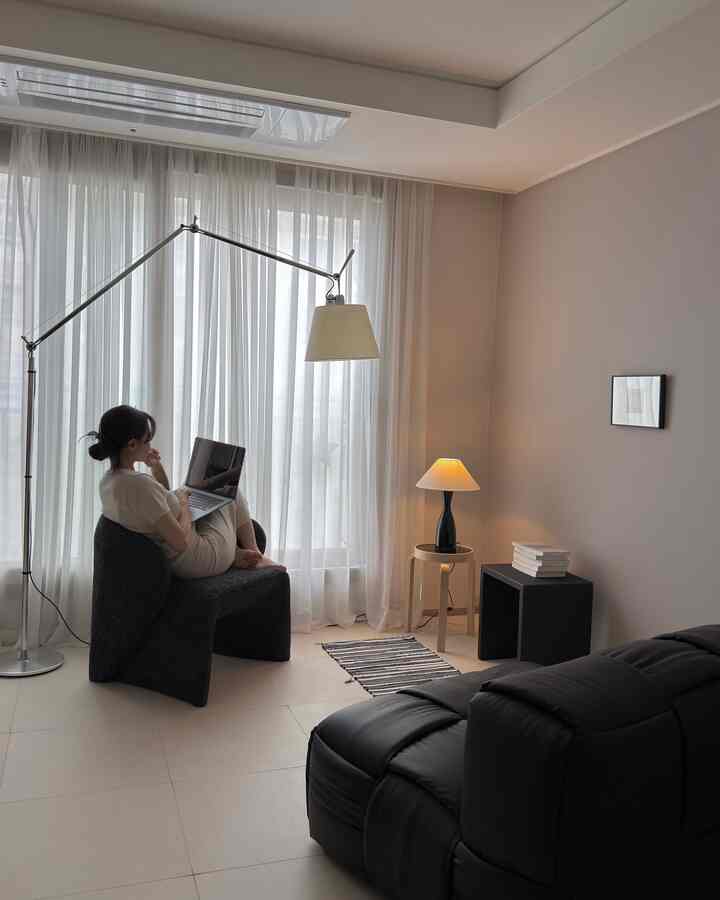 明るい白とダークブラウンのトーンのモダンなリビング、黒い大型ソファとスツール、ナチュラルライトが入るカーテンのある空間