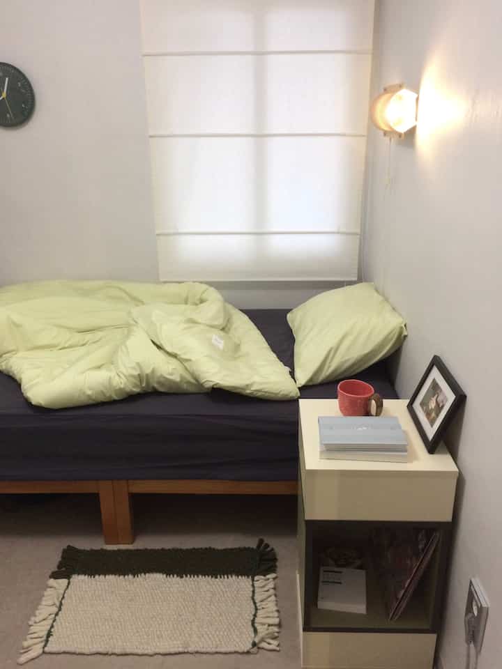 白い壁とネイビーベッドがあるコンパクトなワンルームの寝室、ナイトスタンドに照明と時計が置かれたシンプルな空間