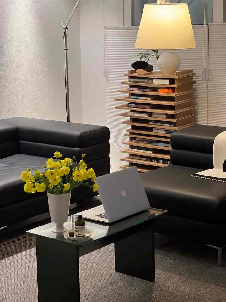 Modern living room with black leather sofa, yellow flowers in a vase, wooden bookshelf, and warm ambient lighting creating cozy atmosphere
