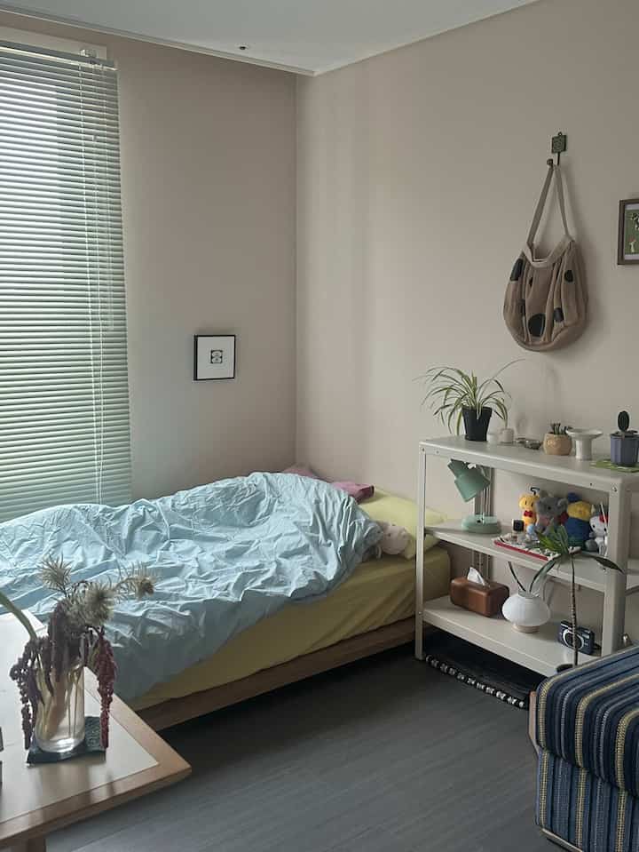 白い壁とブラウンのベッドフレームがある小さな寝室。ミントグリーンのランプや複数の植物が配置されたミニマルで落ち着いた雰囲気のあるインテリア