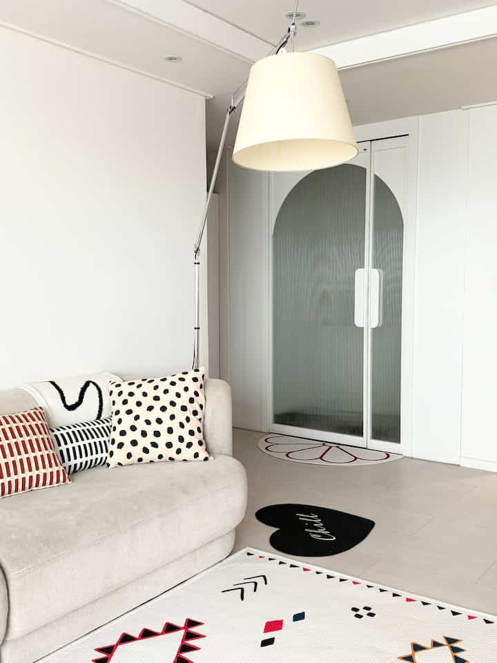 ベージュと白のトーンのリビング、ベージュのソファに柄のクッション、フロアラグ、フロアランプ、アーチ形のガラスドアが配置されたシンプルでモダンなインテリア