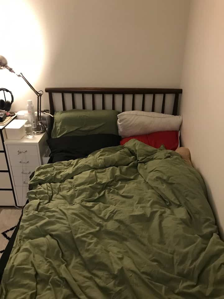 グリーンの寝具とダークブラウンの木製ベッドフレームがある狭い寝室、白い壁と引き出しが配置された空間