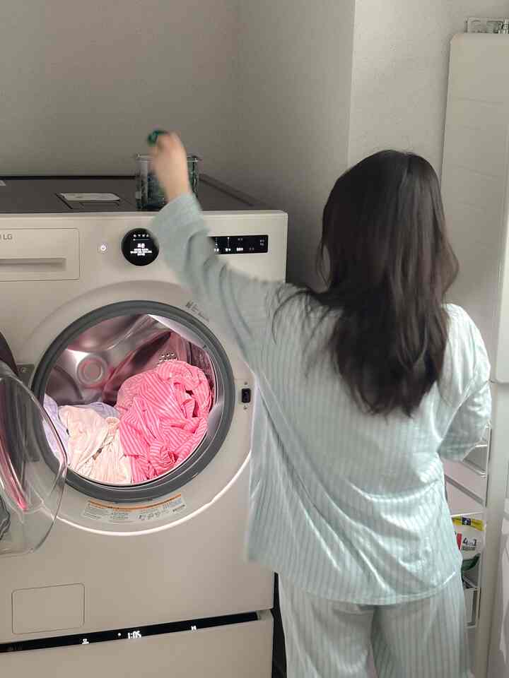 白を基調とした洗濯室で、前面が開いたLGのコンボ洗濯機にピンクの衣類が入っており、使用者が洗剤カプセルを投入している場面