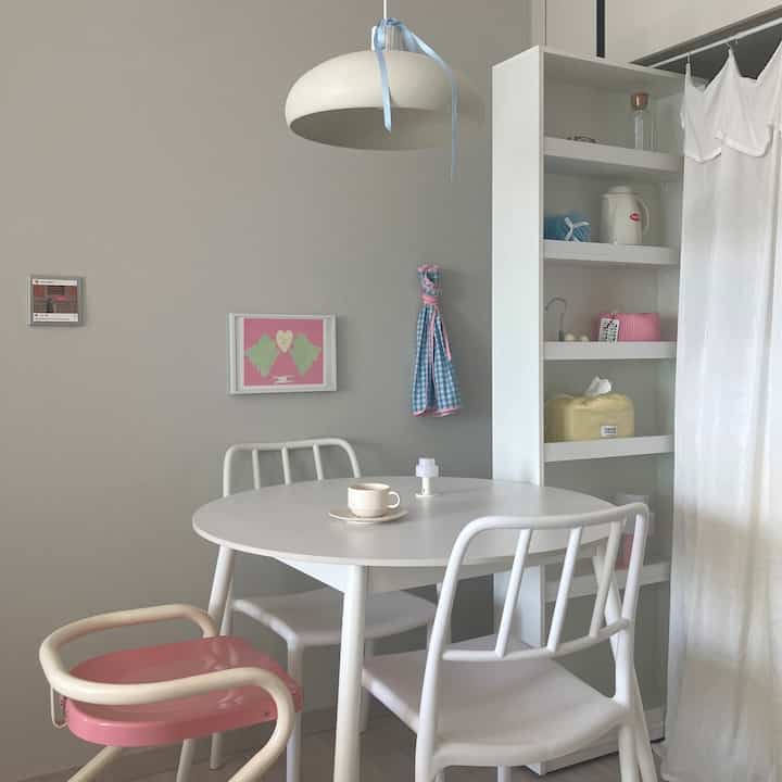 白とパステル調のキッチン、丸いダイニングテーブルと白い椅子、棚が配置され、落ち着いた雰囲気のあるインテリア